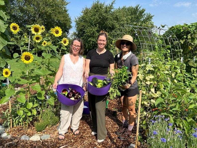 Three women standing in a garden with harvest baskets full of produce.